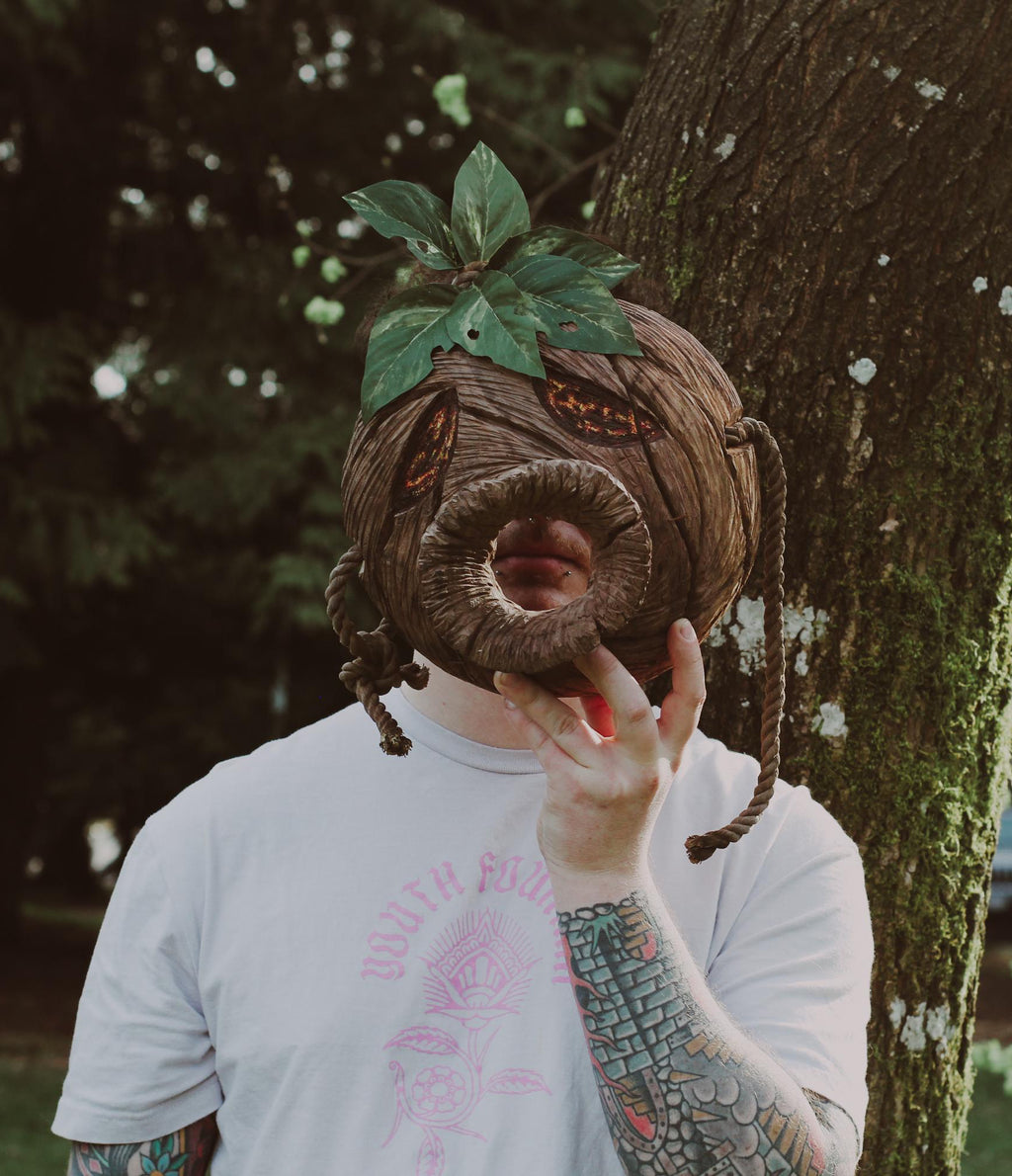 Person with a large wooden head mask holding it to their face, standing next to a tree in an outdoor setting.