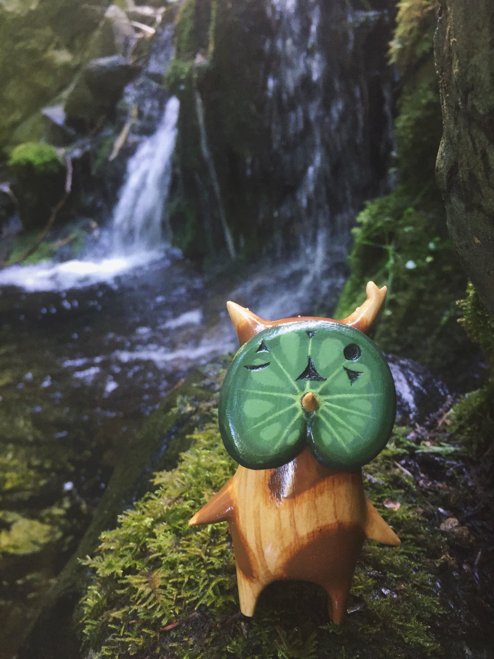 Wooden korok creature with a green leaf face on a mossy rock with a waterfall in the background
