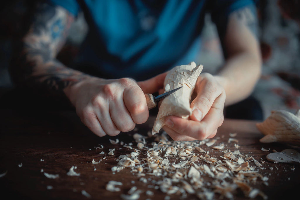 Person carving a wooden figurine with a chisel on a wooden surface