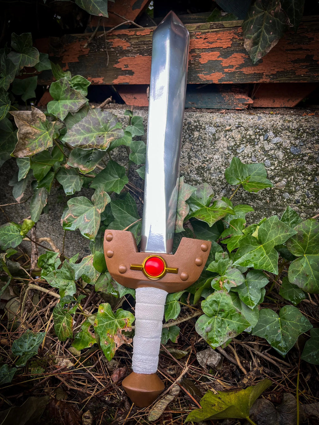 Wooden Kokiri sword with a brown handle and red gem on a stone surface with green leaves.